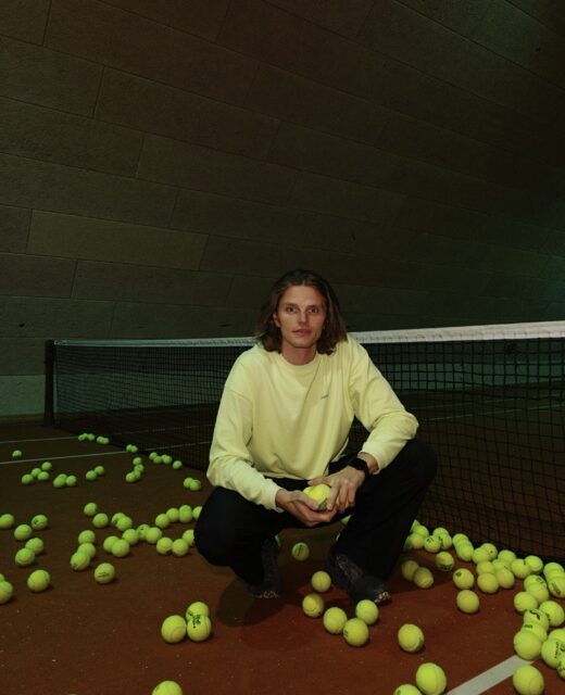 En person sidder foran et net på en indendørs tennisbane, omgivet af mange gule tennisbolde, mens de holder én bold i hænderne.